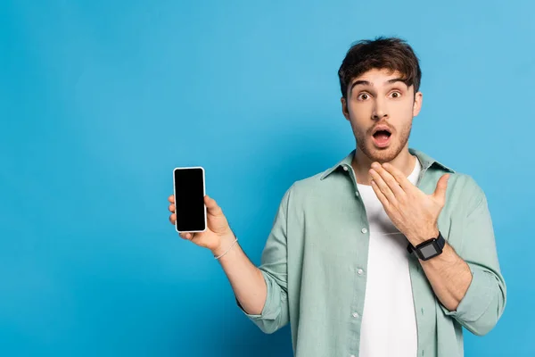 Schockierter junger Mann zeigt Smartphone mit leerem Bildschirm auf blau — Stockfoto