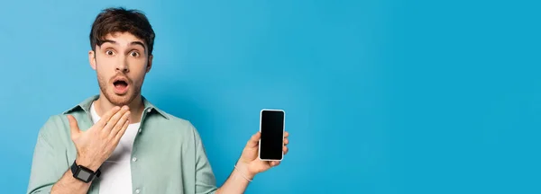 Horizontales Bild eines schockierten jungen Mannes zeigt Smartphone mit leerem Bildschirm auf blauem Hintergrund — Stockfoto