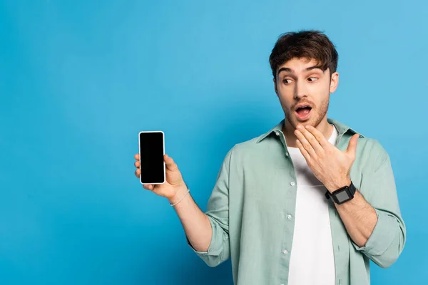 Shocked young man holding hand near mouth while showing smartphone with blank screen on blue — Stock Photo