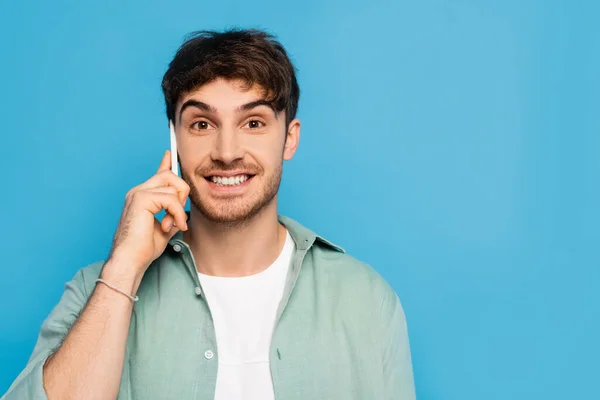 Happy young man talking on smartphone while looking at camera isolated on blue — Stock Photo