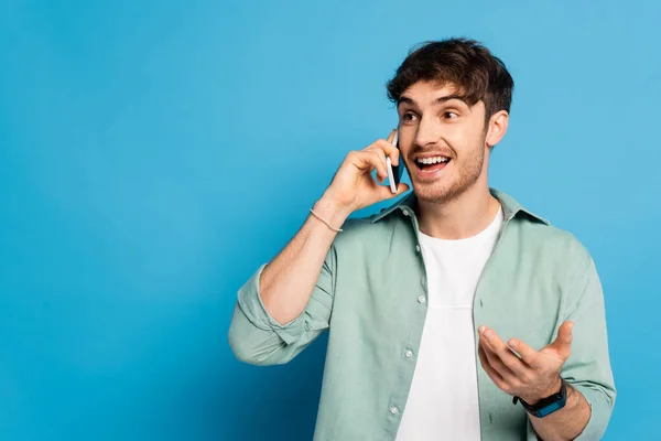 Cheerful young man talking on smartphone while standing with open arm on blue — Stock Photo