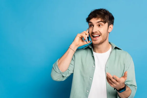 Joyeux jeune homme parlant sur smartphone tout en se tenant debout avec le bras ouvert sur bleu — Photo de stock