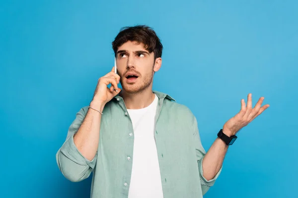 Joven desanimado mostrando gesto de encogimiento mientras habla en el teléfono inteligente en azul - foto de stock