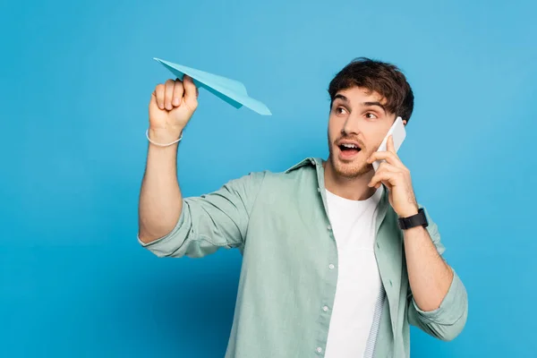 Heureux jeune homme jetant avion en papier tout en parlant sur smartphone sur bleu — Photo de stock