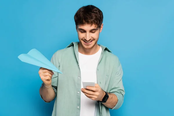 Happy young man chatting on smartphone while holding paper plane on blue — Stock Photo