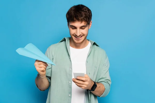 Feliz joven chateando en el teléfono inteligente mientras sostiene el avión de papel en azul - foto de stock