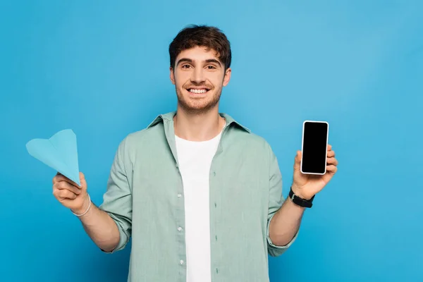 Jovem feliz mostrando smartphone com tela em branco e avião de papel em azul — Fotografia de Stock