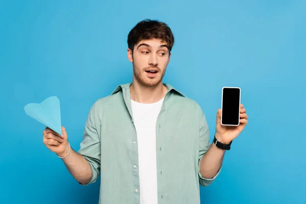 Pensativo joven sosteniendo teléfono inteligente con pantalla en blanco y avión de papel en azul - foto de stock