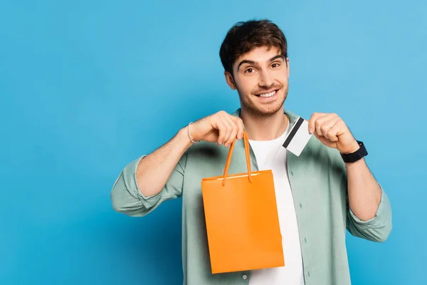 Alegre joven sosteniendo bolsa de compras y tarjeta de crédito en azul - foto de stock