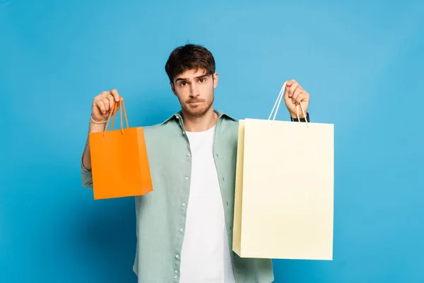 Nachdenklicher junger Mann zeigt Einkaufstüten auf blauem Grund — Stockfoto