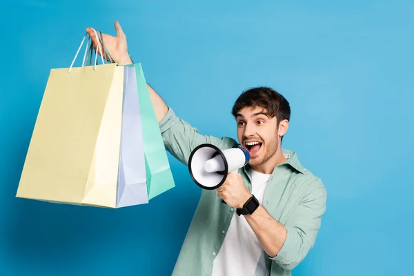 Excité jeune homme criant dans mégaphone tout en tenant des sacs à provisions dans la main levée sur bleu — Photo de stock