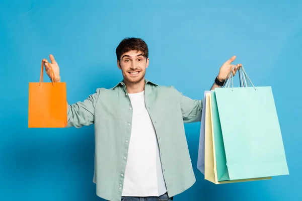 Glücklicher junger Mann hält bunte Papiertüten in der Hand, während er in die Kamera auf blau blickt — Stockfoto