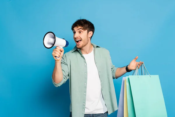 Cheerful young man screaming in megaphone while holding shopping bags on blue — Stock Photo