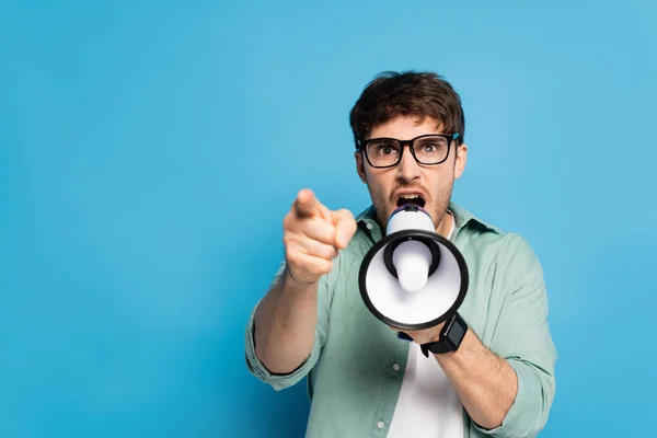 Jovem agressivo gritando em megafone e apontando com o dedo no azul — Fotografia de Stock