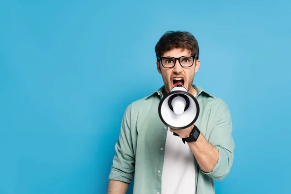 Wütender junger Mann schreit in Megafon, während er auf blauem Grund in die Kamera schaut — Stockfoto