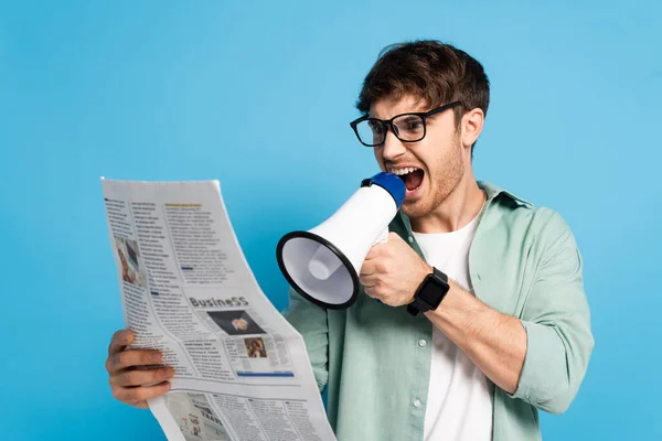Сердитий молодий чоловік кричить в мегафоні під час читання газети на синьому — стокове фото