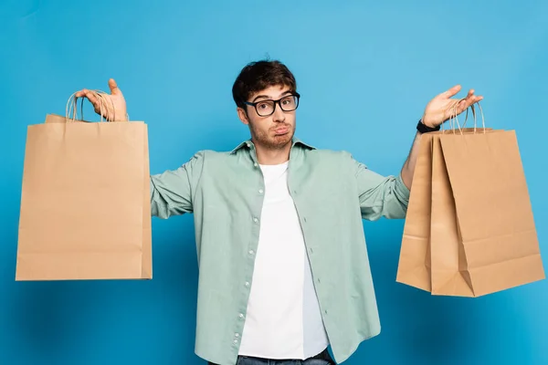 Confundido joven sosteniendo bolsas de compras mientras mira la cámara en azul - foto de stock