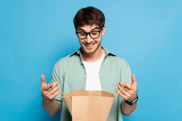 Überraschter junger Mann schaut in Einkaufstasche auf blauem Grund — Stockfoto