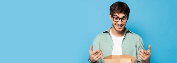 Horizontal crop of surprised young man looking into shopping bag on blue — Stock Photo