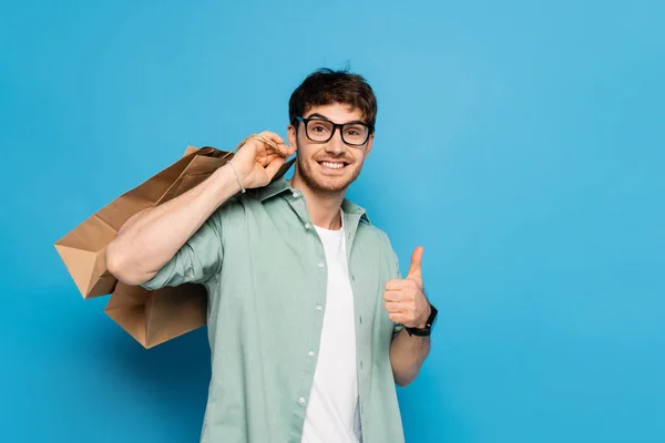 Glücklicher junger Mann zeigt Daumen nach oben, während er Einkaufstüten auf blauem Grund trägt — Stockfoto