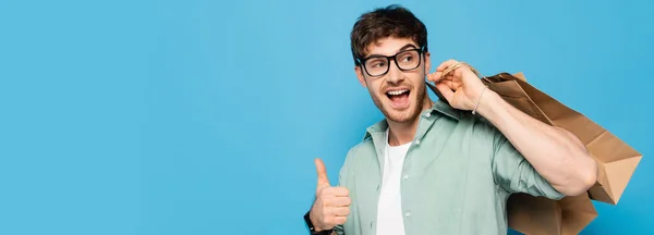 Website-Header eines glücklichen jungen Mannes zeigt Daumen nach oben, während er Einkaufstüten isoliert auf blauem Grund trägt — Stockfoto