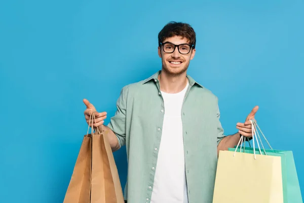 Feliz joven mirando a la cámara mientras sostiene bolsas de compras en azul - foto de stock