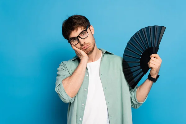 Exhausted young man touching face while holding hand fan on blue — Stock Photo