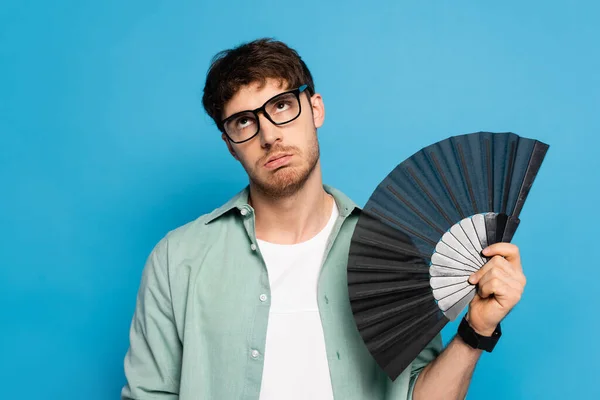 Exhausted young man holding hand fan while suffering from summer heat on blue — Stock Photo