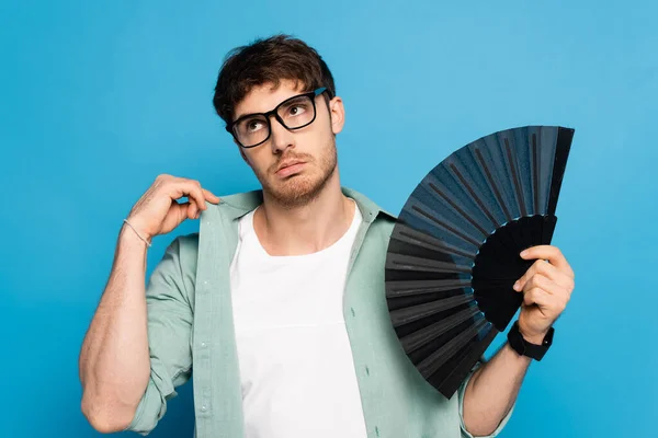 Scontento giovane uomo toccando camicia mentre tiene ventilatore mano sul blu — Foto stock