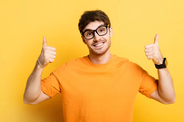 Cheerful young man smiling at camera and showing thumbs up on yellow — Stock Photo