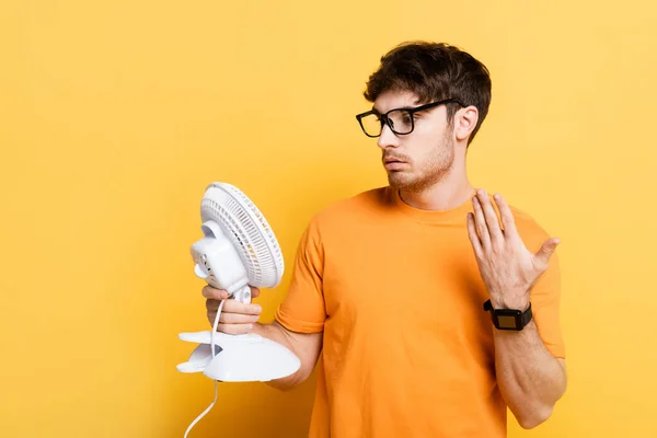 Scontento giovane uomo agitando mano mentre tiene ventilatore elettrico sul giallo — Foto stock