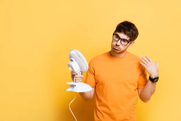 Homem descontente segurando ventilador elétrico e acenando mão no amarelo — Fotografia de Stock
