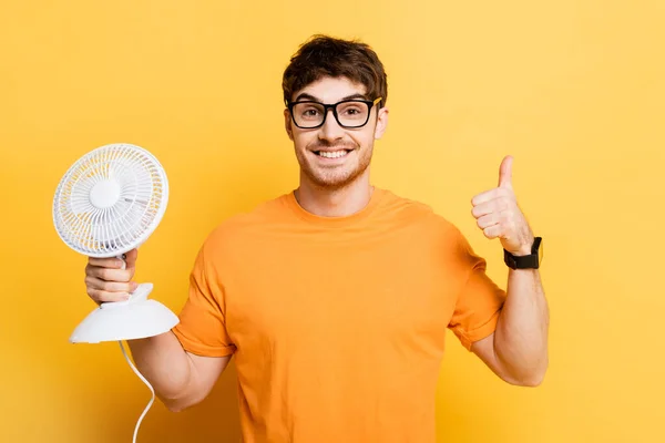 Feliz joven sosteniendo ventilador eléctrico y mostrando los pulgares hacia arriba en amarillo - foto de stock