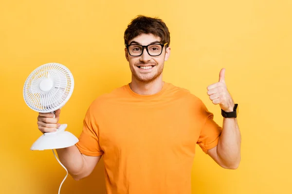 Jovem feliz segurando ventilador elétrico e mostrando polegares para cima no amarelo — Fotografia de Stock