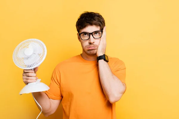 Esausto giovane uomo toccare il viso mentre tiene ventilatore elettrico su giallo — Foto stock