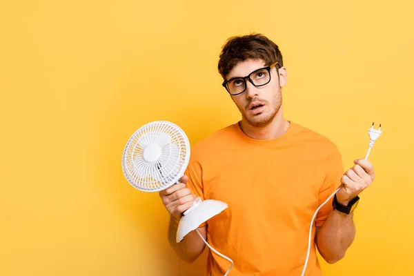 Истощенный молодой человек держит электрический вентилятор и смотрит вверх, страдая от жары на желтый — стоковое фото