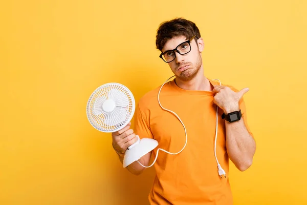 Hombre disgustado sosteniendo ventilador eléctrico mientras que sufre de calor en amarillo - foto de stock