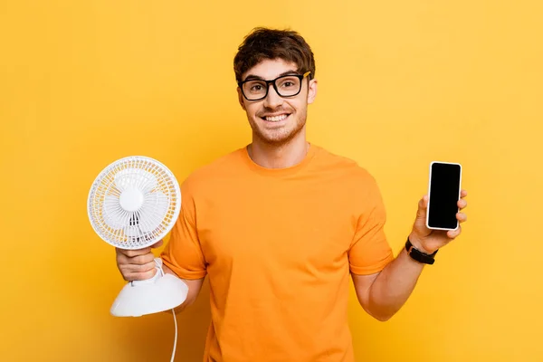 Joven feliz sosteniendo ventilador eléctrico y teléfono inteligente con pantalla en blanco en amarillo - foto de stock