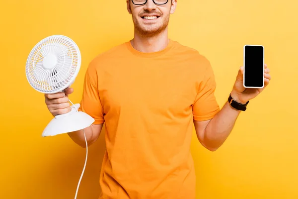 Vista ritagliata dell'uomo sorridente che tiene smartphone con schermo vuoto e ventilatore elettrico sul giallo — Foto stock