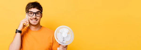 Panoramic shot of cheerful young man talking on smartphone while holding electric fan isolated on yellow — Stock Photo