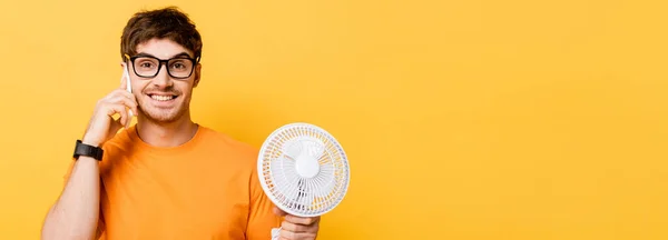 Plano panorámico de joven alegre hablando en el teléfono inteligente mientras sostiene ventilador eléctrico aislado en amarillo - foto de stock