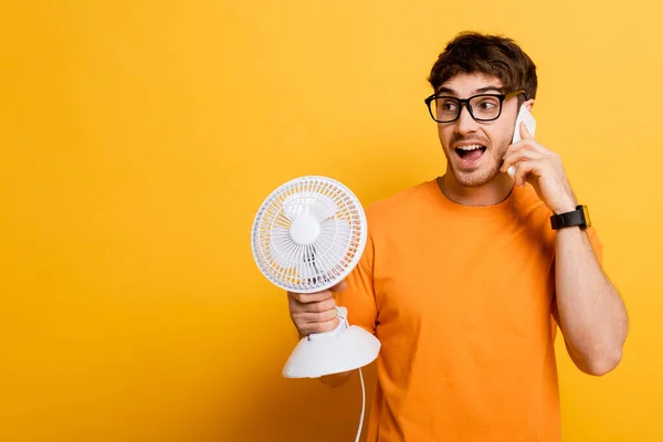 Jovem animado falando no smartphone enquanto segurando ventilador elétrico no amarelo — Fotografia de Stock