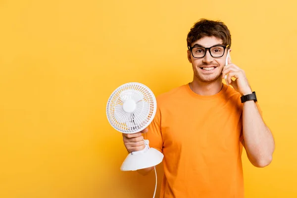 Fröhlicher Mann spricht auf Smartphone, während er einen elektrischen Ventilator auf gelb hält — Stockfoto