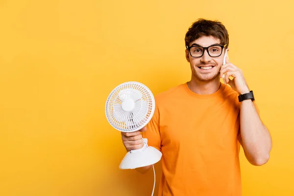 Homem alegre falando no smartphone enquanto segurando ventilador elétrico no amarelo — Fotografia de Stock