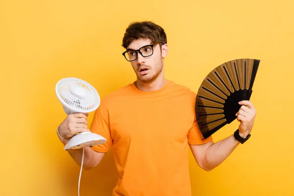 Homme mécontent tenant ventilateurs électriques et de la main tout en souffrant de chaleur sur jaune — Photo de stock