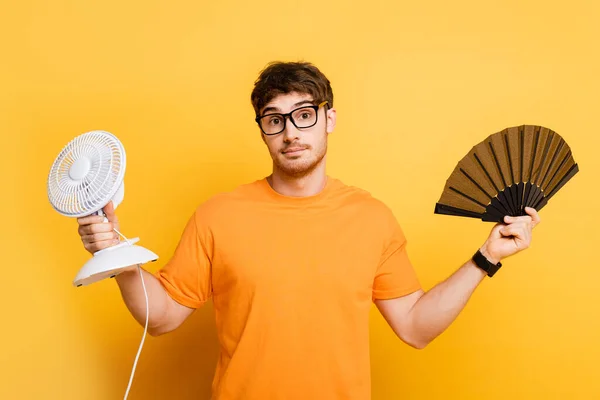 Discouraged young man holding electric and hand fans while looking at camera on yellow — Stock Photo