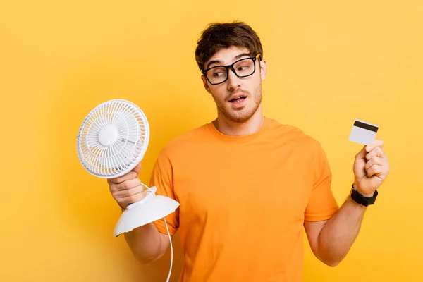 Jovem pensativo segurando ventilador elétrico e cartão de crédito no amarelo — Fotografia de Stock