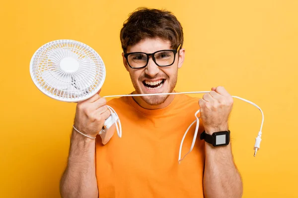 Uomo arrabbiato in occhiali tenendo rotto ventilatore elettrico su giallo — Foto stock