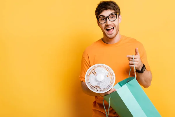 Sorpreso giovane uomo in occhiali tenendo borsa della spesa con nuovo ventilatore elettrico su giallo — Foto stock