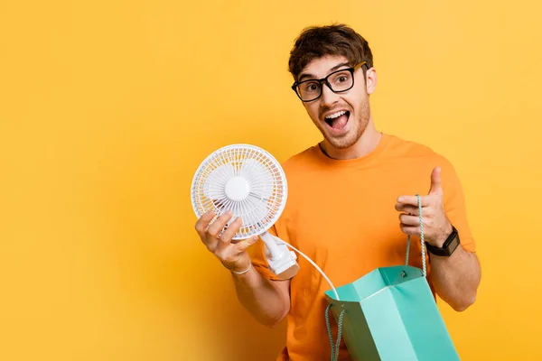 Aufgeregter Mann mit Brille hält Einkaufstasche mit neuem Elektroventilator auf gelb — Stockfoto